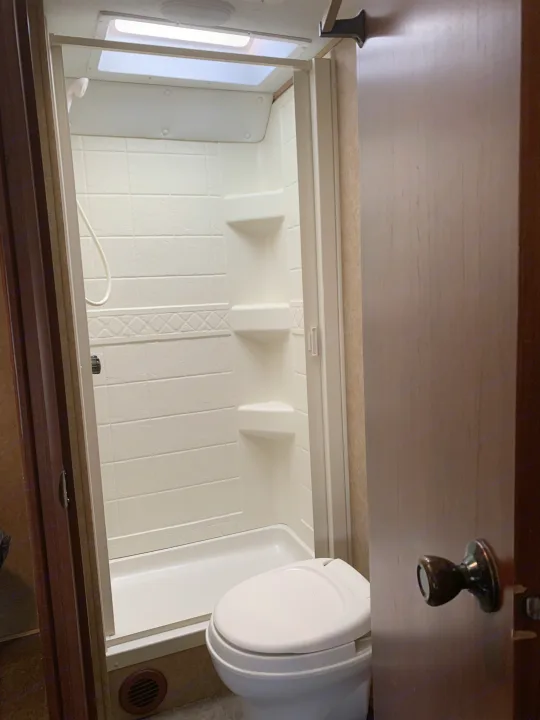 Inside an RV rental, showing a compact white bathroom with a shower stall, toilet, and wooden door on the right.. Forest River Sunseeker 2014