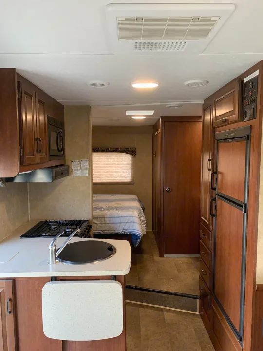 This RV rental features a full kitchen with a stove, sink, and fridge, plus a bedroom visible in the background. A compact mobile living space.. Forest River Sunseeker 2014