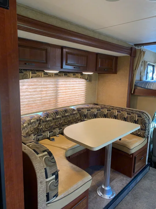 Cozy RV rental dinette with U-shaped patterned seating, light table, dark wood cabinets, and window blinds. Bunk bed visible.. Forest River Sunseeker 2014