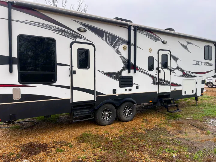 White Fuzion RV rental, a fifth-wheel toy hauler, with bold graphics, dual axles, and two entry doors on gravel.. Keystone Fuzion 2015
