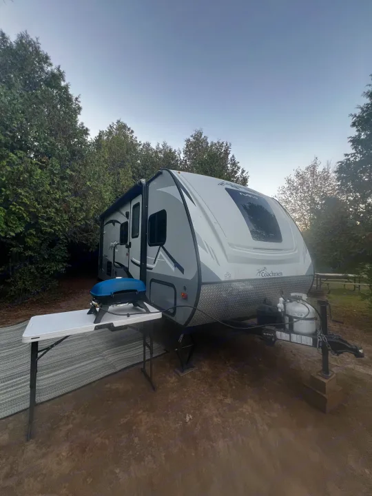 Our RV rental, a Coachmen Apex Nano travel trailer, parked at a quiet, tree-lined campsite with a grill.. Apex Nano 193 BHS 2020