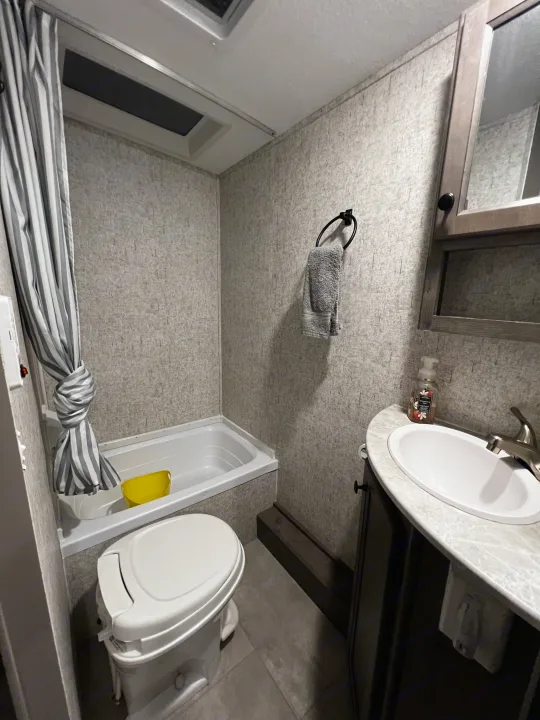 Inside an RV rental, this compact bathroom features a white toilet, small tub with striped curtain, and a vanity sink.. Apex Nano 193 BHS 2020