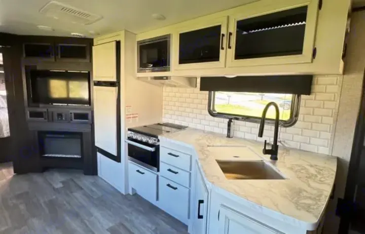 White & grey RV rental interior: modern kitchen with subway tile, sink, stove, microwave; plus TV & fireplace.. Jayco White Hawk 2023