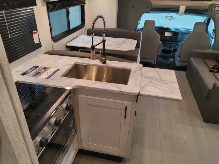 Modern RV rental interior: kitchen with a large sink, dinette, and sofa. Driver's cabin visible in background.. East To West Entrada 2025