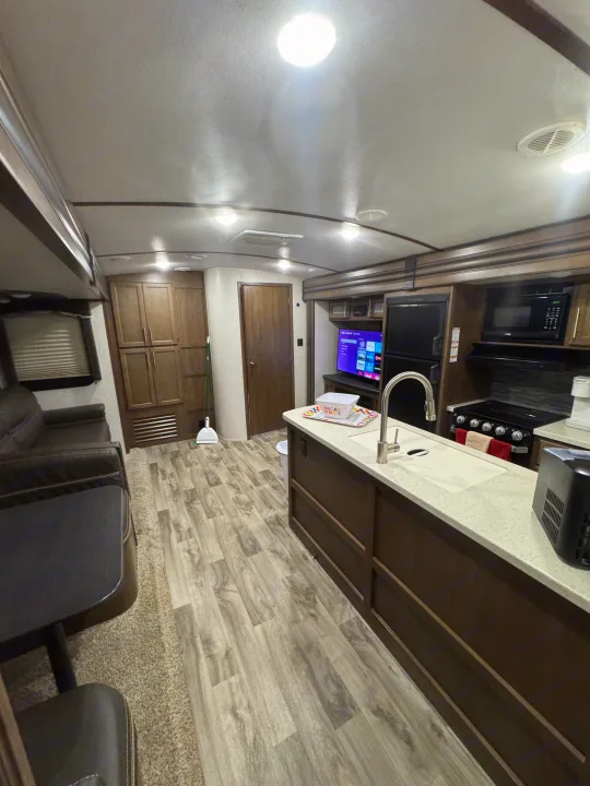 Inside a modern RV rental: living area with dark sofa & table, full kitchen with sink, stove, microwave, and wood floors. Well-lit.. Keystone RV Cougar 2019