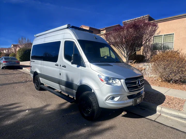 A silver Mercedes-Benz Sprinter camper van for RV rental, parked on a sunny residential street.. Thor Sanctuary 19P 2025