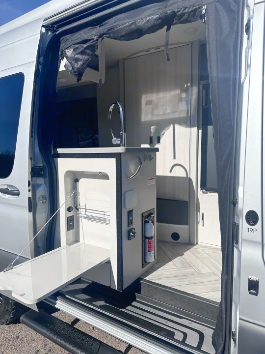 RV rental's side door reveals a compact kitchen with a sink, fold-out counter, and fire extinguisher for safety.. Thor Sanctuary 19P 2025