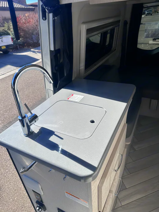 RV rental: A bright, compact galley with a chrome faucet, light grey counter, and wood cabinets, viewed from the open door.. Thor Sanctuary 19P 2025