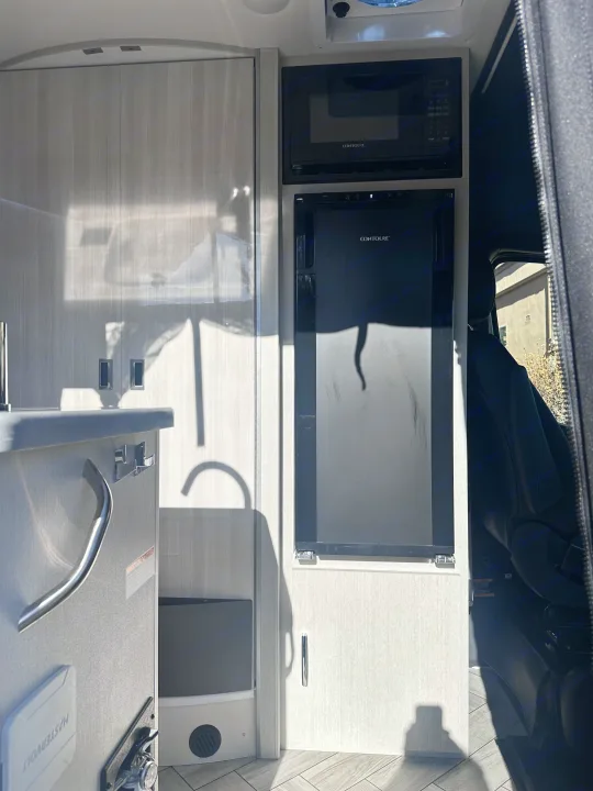 Inside an RV rental: bright galley with a chrome faucet, grey speckled countertop, and covered sink. Side door open to street view.. Thor Sanctuary 19P 2025