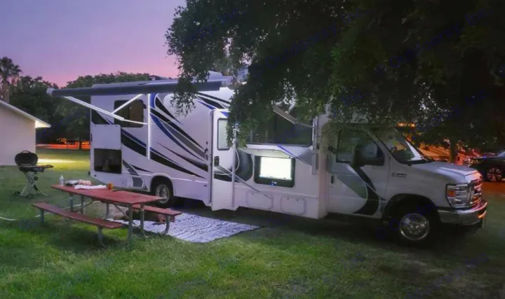 Evening RV rental camping: A white RV with an extended awning, picnic table, grill, and outdoor TV under a twilight sky.. Thor Four Winds 2021