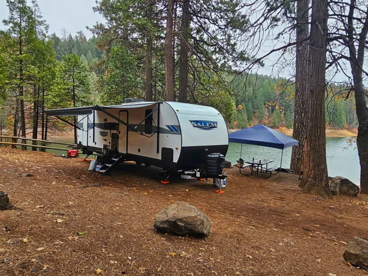 Lakeside RV rental campsite with a white travel trailer, blue canopy, and picnic table nestled in a forest setting.. Forest River Salem 2024