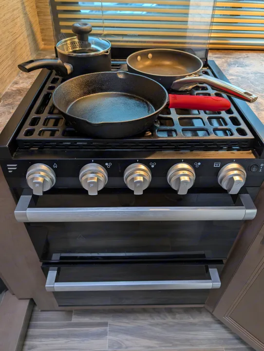 RV rental kitchen featuring a compact stove/oven, with a cast iron skillet, saucepan, and frying pan on the grates.. Thor Four Winds 2021