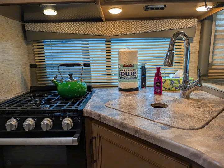 An RV rental kitchen/galley shows a green kettle on a gas stove, a sink with chrome faucet, and countertop items.. Thor Four Winds 2021