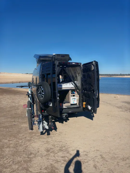 Black camper van RV rental with bike, parked by a scenic lake. Open rear doors reveal custom interior, ready for adventure!. Winnebago Winnebago 2024