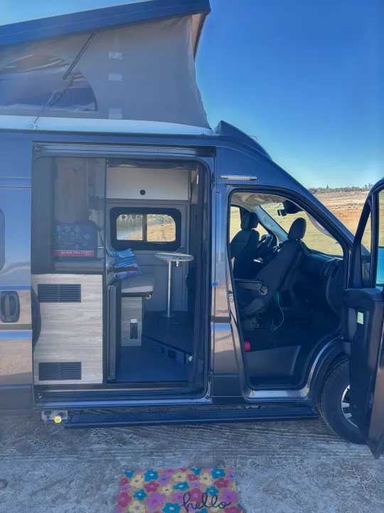 Explore inside this gray pop-top camper van RV rental! Side door open, showing inviting interior & outdoor scenery.. Winnebago Winnebago 2024