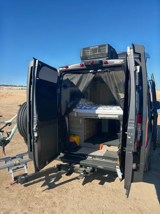An RV rental’s open rear reveals a bed, storage, and mesh screen. A bike and spare tire are mounted on the exterior rack.. Winnebago Winnebago 2024