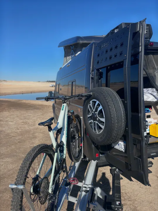 Adventure awaits! This RV rental features a pop-top van, bike rack with mountain bike, and spare tire, parked by water.. Winnebago Winnebago 2024