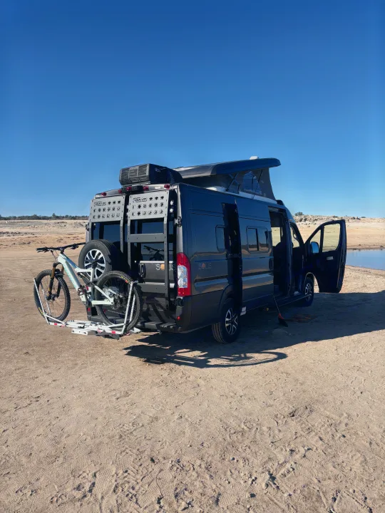 This dark grey RV rental, equipped with a mountain bike rack, is parked by a lake, ready for outdoor adventure.. Winnebago Winnebago 2024