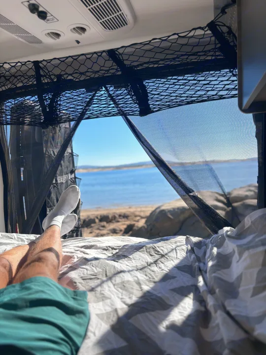 Inside an RV rental, a person's legs are stretched out on a bed, gazing at a serene lake through mesh screens.. Winnebago Winnebago 2024