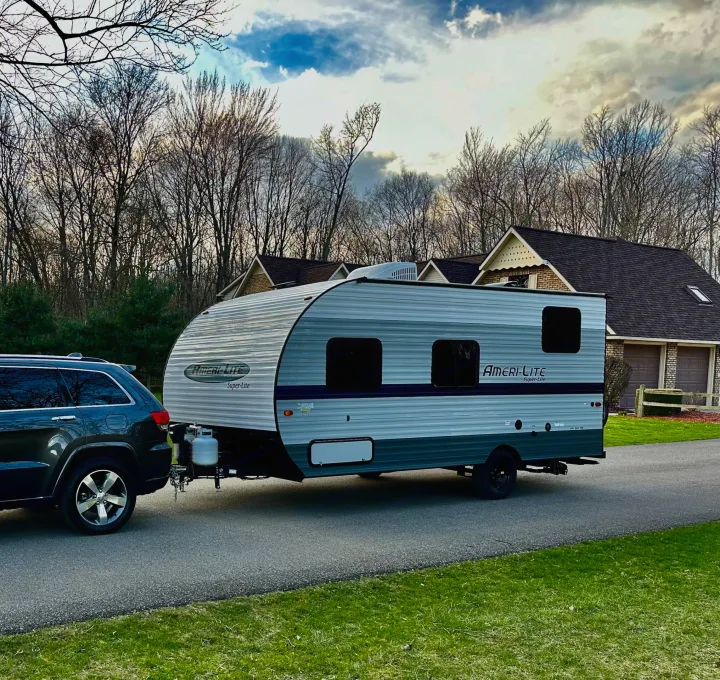 Dark SUV towing a light grey Ameri-Lite travel trailer on a paved driveway, likely an RV rental ready to go.. Gulf Stream Ameri-Lite Super Lite 2021