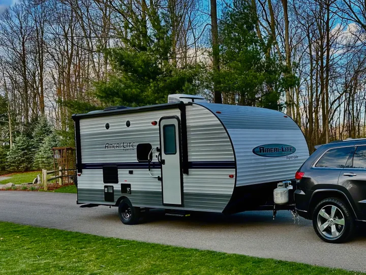 An Ameri-Lite Super-Lite travel trailer is hitched to an SUV on a driveway, ready for RV rental adventures through the bare trees.. Gulf Stream Ameri-Lite Super Lite 2021