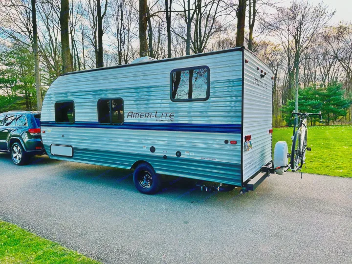 Side view of a light grey Ameri-Lite travel trailer (RV rental) hitched to an SUV, with a bicycle on its rear rack.. Gulf Stream Ameri-Lite Super Lite 2021