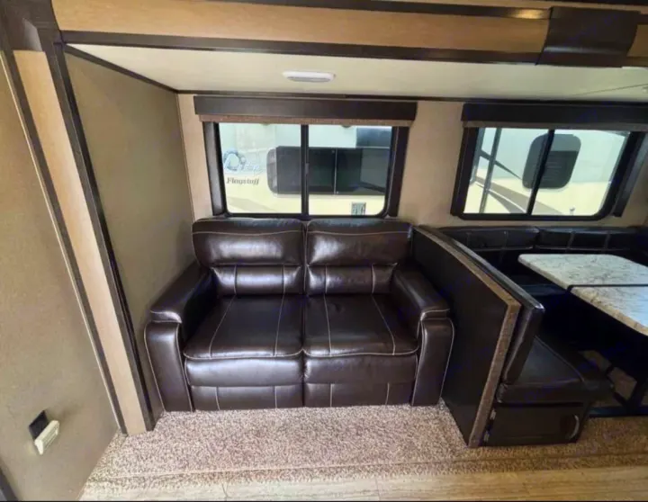 This RV rental interior offers a brown sofa, a dinette with bench seating, and windows, all on a carpeted floor. Ideal for travel.. Grand Design Imagine 2018
