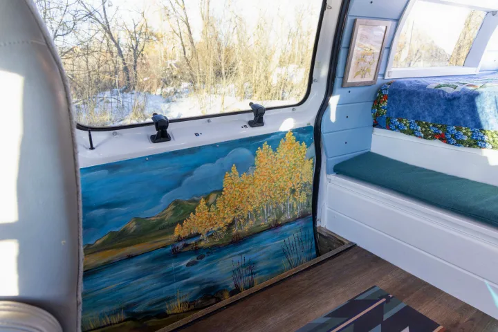 This RV rental's custom campervan interior features a scenic painted wall, cozy bench, bed, and snowy window view.. Ford Econoline 150 2009