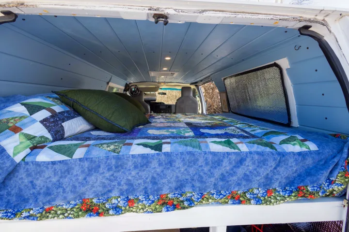 Inside an RV rental: a comfy bed with a quilted blanket & blue wood-paneled ceiling in a custom camper van.. Ford Econoline 150 2009