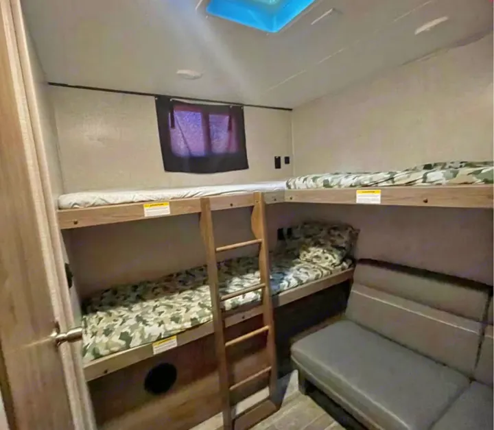 Inside an RV rental: bunk beds with camo bedding, a small ladder, window, and a gray couch. Compact sleeping and lounging space.. Crossroads RV Zinger 2021