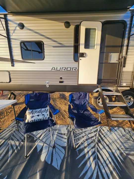 An RV rental parked with two camping chairs and an outdoor mat, ready for adventure at a sunny campsite.. Forest River Aurora Aurora 2024
