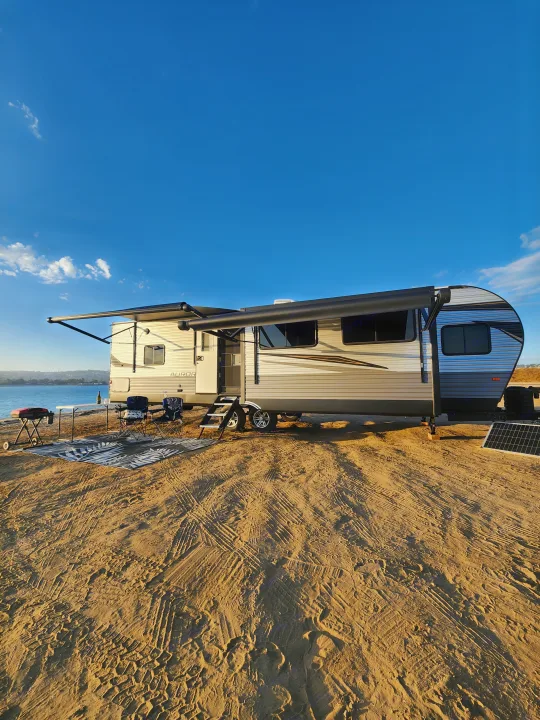Enjoy your RV rental! This travel trailer is set up with awnings, grill, and chairs on a sandy beach by the water.. Forest River Aurora Aurora 2024