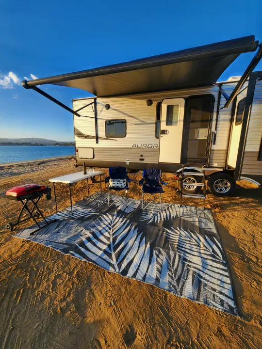 Scenic RV rental on a sandy beach. Awning extended over an outdoor setup with chairs, table, grill, and patterned mat.. Forest River Aurora Aurora 2024