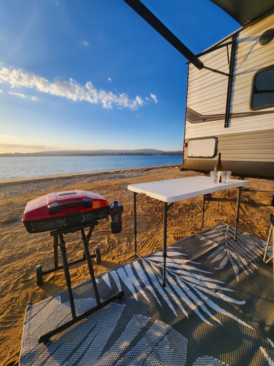 RV rental escape: waterfront camping with a travel trailer, an outdoor grill, table, and mat setup on the sand.. Forest River Aurora Aurora 2024