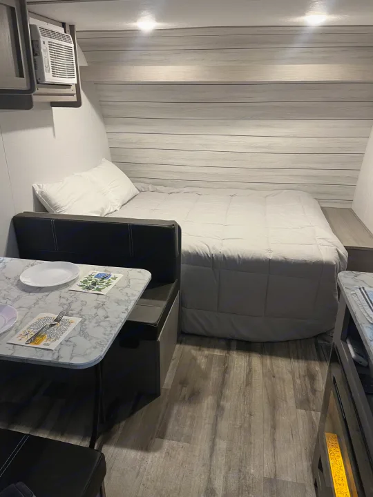 This rv rental features a cozy interior with a light gray bed, a marble-top dining booth, and an overhead AC unit.. Dutchmen Coleman Lantern LT 17B 2024