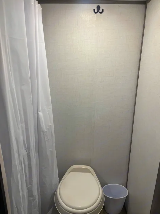 A compact rv rental bathroom, showcasing a white toilet, shower curtain, and practical wall hooks on textured gray walls.. Dutchmen Coleman Lantern LT 17B 2024