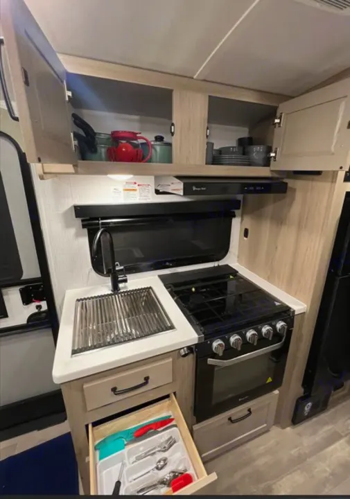 RV rental kitchen featuring a compact stove, sink with drainer, open overhead cabinets with cookware, and an open utensil drawer.. Forest River Rockwood Geo Pro 2024