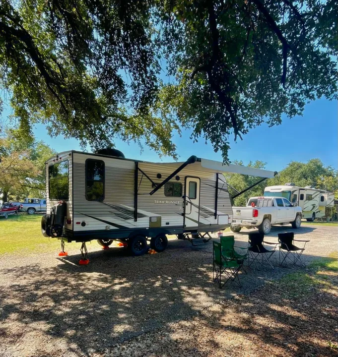 RV rental fun: "Trail Runner" travel trailer parked under shade trees, awning extended, with camping chairs.. Heartland RVs Trail Runner SLE 2019
