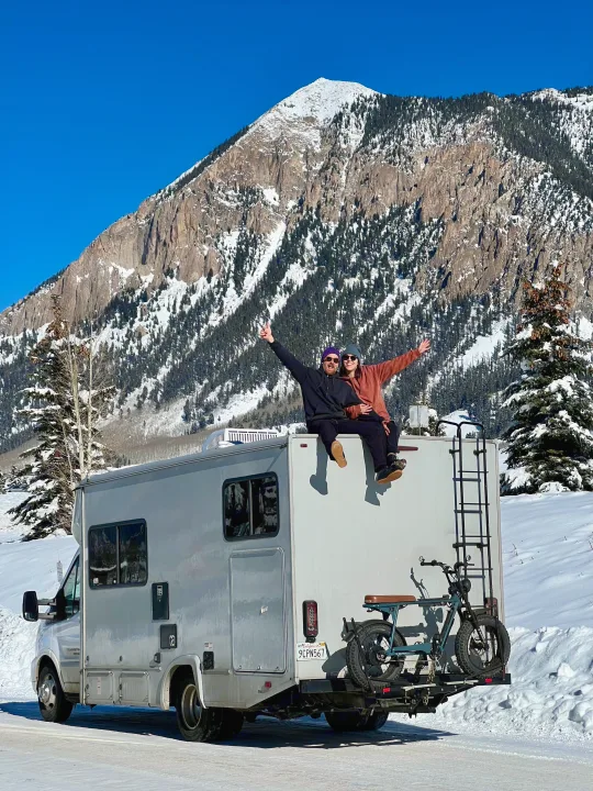 A couple sits atop their RV rental, enjoying a stunning snowy mountain view. Winter travel adventure!. Coachmen Crosstrail 20XG 2022