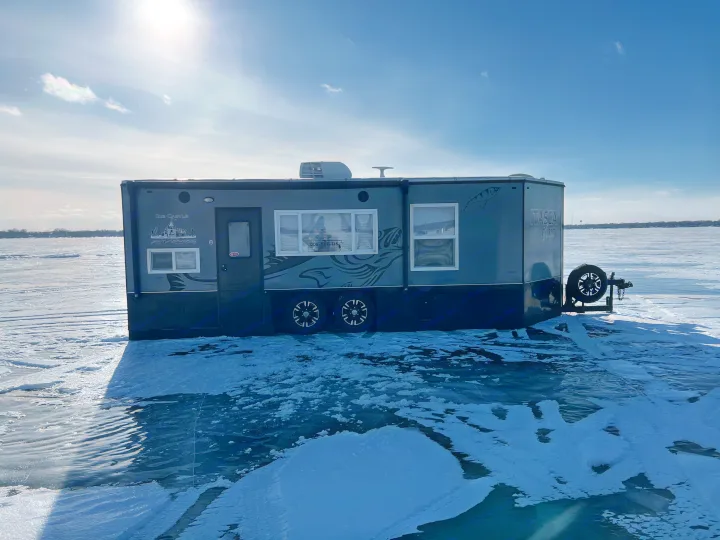 This Ice Castle RV rental, adapted for ice fishing, sits on a sunny frozen lake. Ideal for winter outdoor adventures.. Ice Castle Fish Houses Toy Hauler 2019