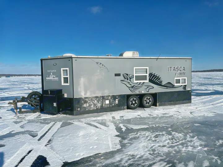 An Itasca Extreme RV rental ice fishing house from Ice Castle, parked on a vast frozen lake with snow under blue sky.. Ice Castle Fish Houses Toy Hauler 2019