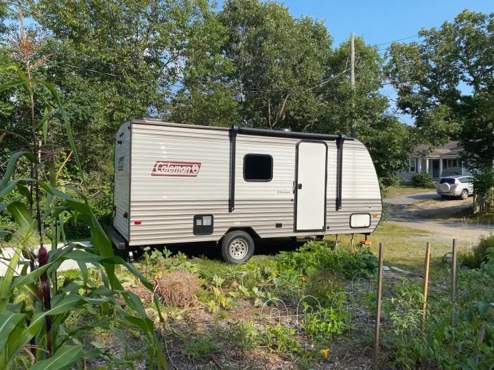 Coleman travel trailer RV rental, set up in a yard next to a vibrant garden with corn plants.. Coleman Lantern 17B 2024