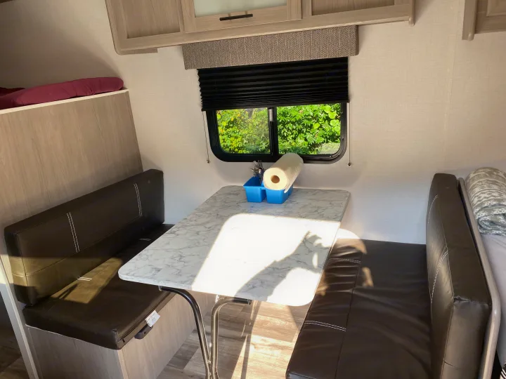 Spacious RV rental dinette featuring a sunny marble-look table, dark benches, and a large window with lush green views.. Coleman Lantern 17B 2024