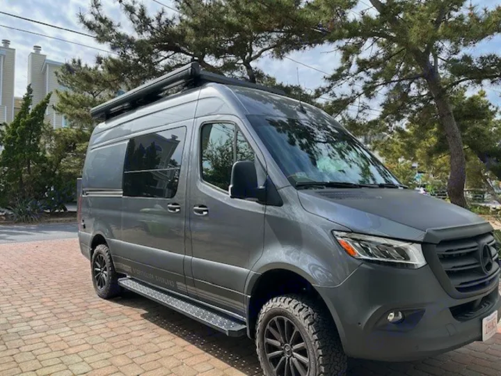 A sleek, dark gray Mercedes Sprinter camper van, equipped with off-road tires and an awning. Perfect for an RV rental.. Storyteller Overland Stealth Mode 2023