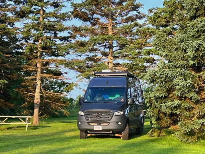 Modern dark gray Sprinter RV rental parked on grass surrounded by tall evergreen trees, ready for your next adventure.. Storyteller Overland Stealth Mode 2023