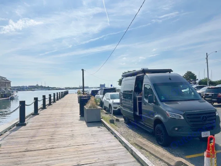 An RV rental, a dark gray Mercedes Sprinter camper van, is parked next to a wooden pier by a harbor.. Storyteller Overland Stealth Mode 2023