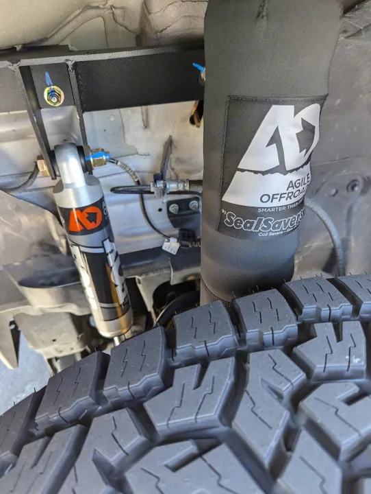 This close-up shows robust off-road suspension components of an RV rental, including FOX shocks & Agile Offroad covers, next to a rugged tire.. Thor Sanctuary 2022