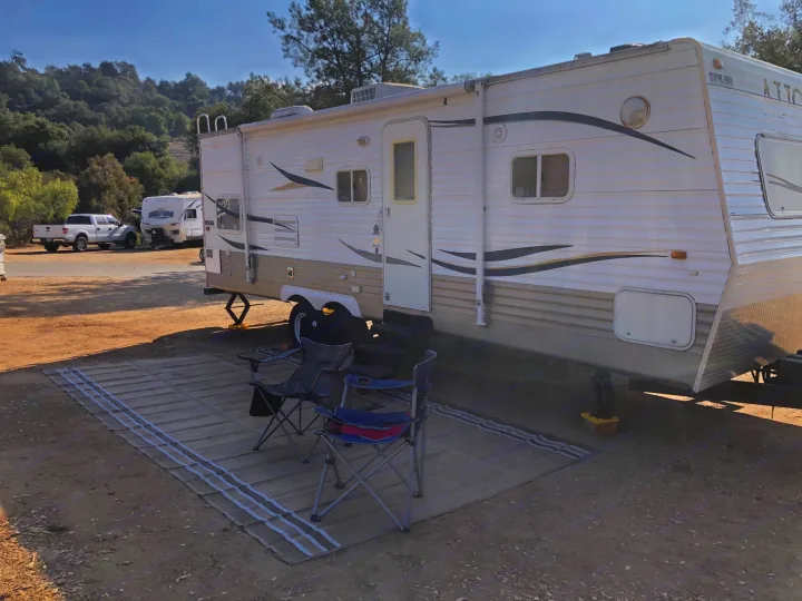 An RV rental travel trailer is parked on a dirt campsite with an outdoor mat and chairs. Another RV & truck are nearby. Enjoying the outdoors.. Aljo Aljo Trailer 2008