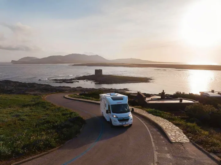 An RV rental drives on a coastal road overlooking a historic island tower, sea, and mountains in the golden light.. Dethleffs JUST T7052 DBM 2023