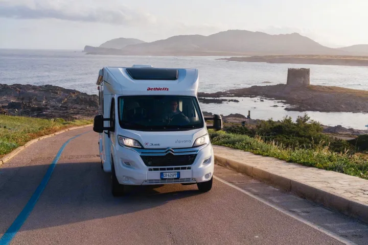 A white Dethleffs motorhome rv rental navigates a coastal road with a blue line, an ancient stone tower visible across the water.. Dethleffs JUST T7052 DBM 2023